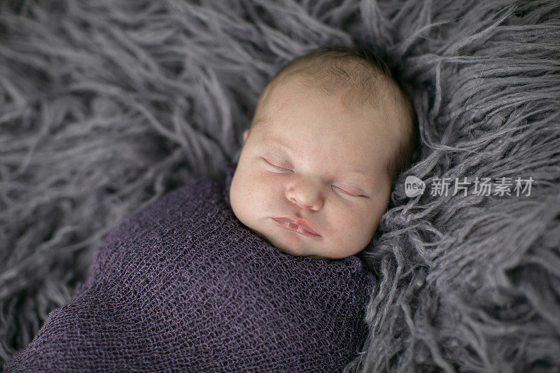 这是一个可爱的新生婴儿的特写镜头，小女孩躺在紫色的毛绒毯子上，全身包裹着