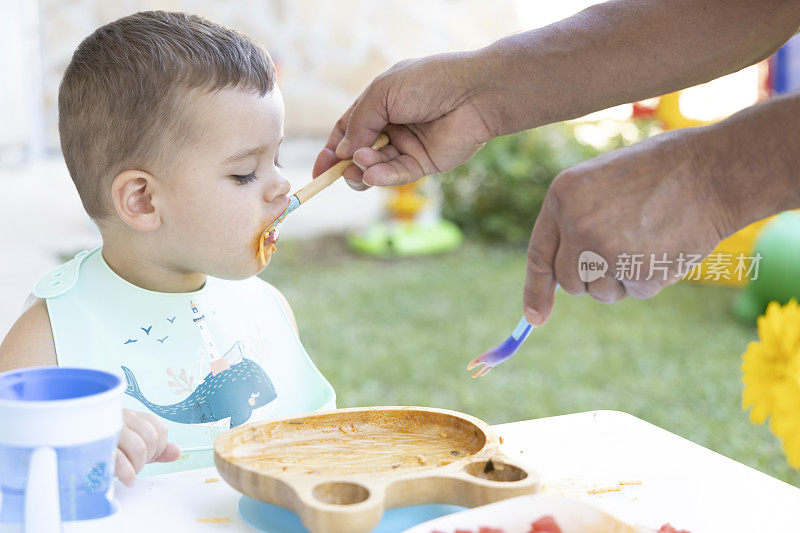 一个可爱的小男孩在他爷爷的帮助下吃饭。一个小男孩坐在花园里的椅子上吃东西。