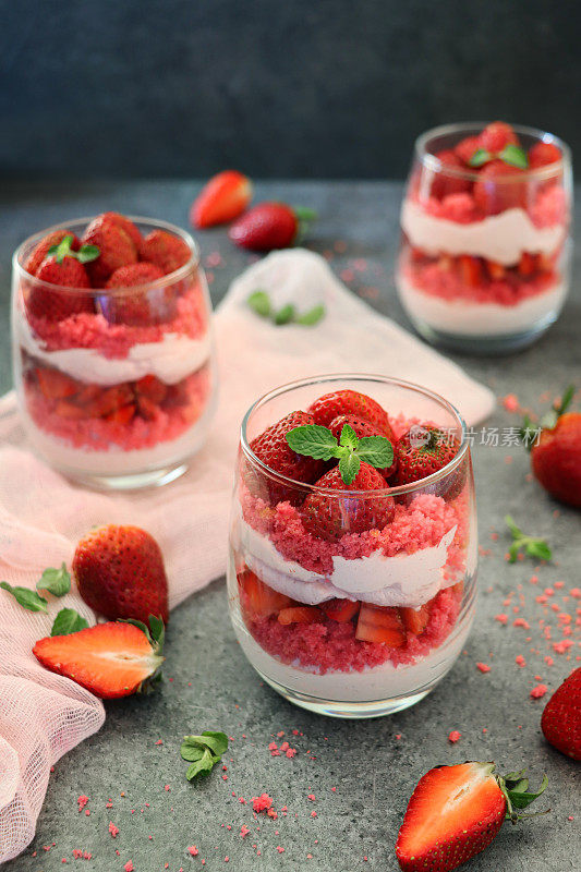 图像眼镜包含自制的草莓慕斯甜点，上面覆盖着整个草莓，奶油慕斯和红丝绒蛋糕屑的分层布丁，周围是蛋糕屑和水果，灰色的表面，高架的视野，聚焦前景