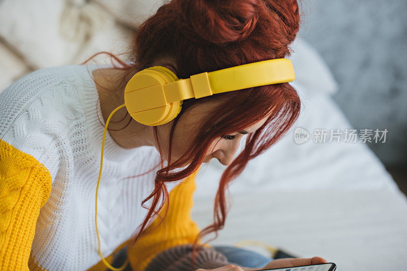 一个红头发的女孩躺在床上，戴着黄色的耳机听音乐。