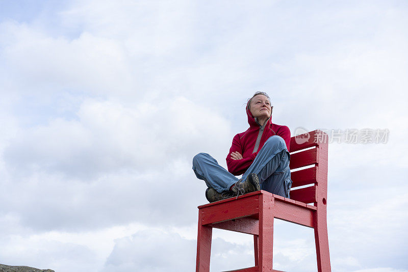 老妇人坐在巨大的红色椅子上，看着冰岛东南角的蝙蝠侠山