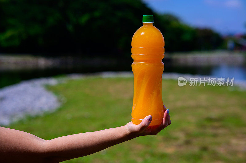 在公园里，女人的手拿着一个大塑料瓶的橙汁。户外