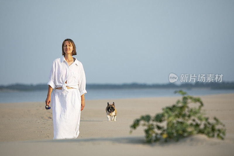 年轻的成年女子和她的法国斗牛犬在海滩上散步