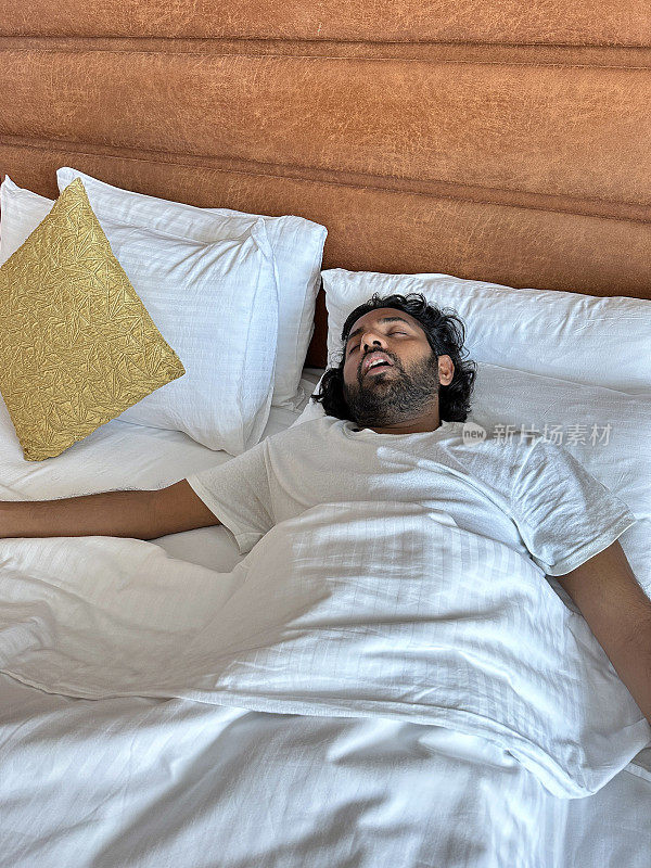 印度男子躺在酒店房间的双人床上的枕头上，鼾声如雷，睡在白色床单下，人造革床头板，抬高视野，专注于前景