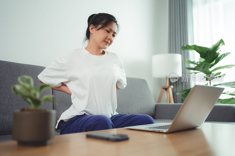 一名亚洲女性坐在家里的沙发上使用笔记本电脑，身体疼痛。