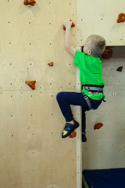 儿童攀岩。穿绿衬衫的金发男孩正在爬墙。抱石类。为安全活动及比赛而设的人造跑道。登山，安全系统，专用鞋