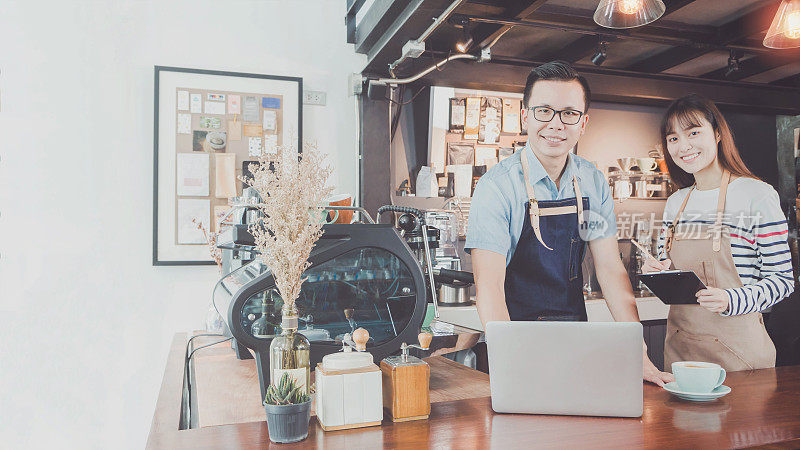 年轻的亚洲男女咖啡师穿着围裙，用手提电脑和咖啡杯微笑着在咖啡厅柜台招待顾客。咖啡店和小型咖啡店的概念。的语气