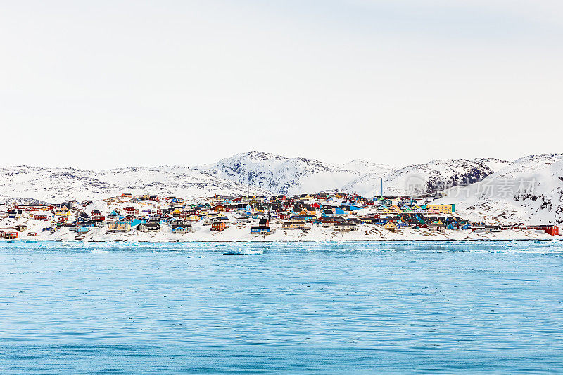 格陵兰岛伊卢利萨特的北极城市全景，色彩斑斓的因纽特人的房子在被雪覆盖的岩石山上，背景是山