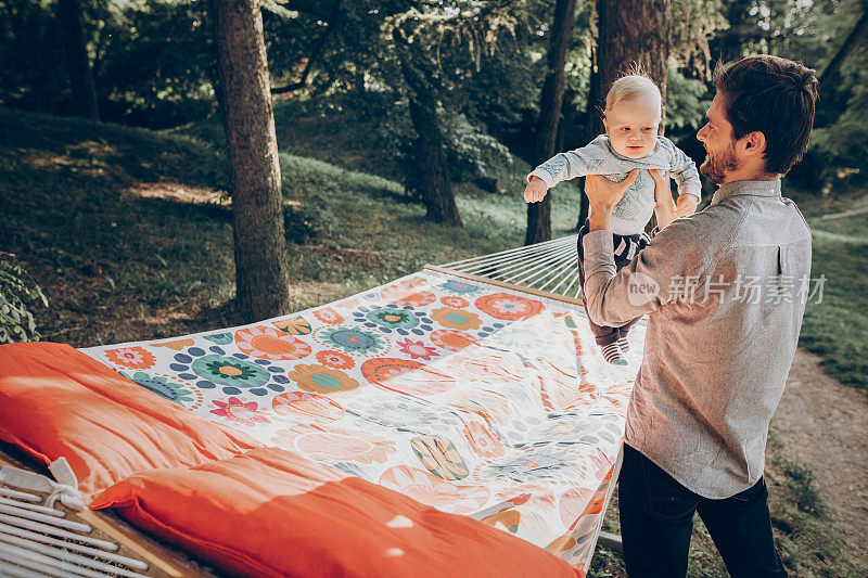 快乐的潮爸爸和他的儿子在公园附近的吊床上玩，而在森林露营，家庭的概念，英俊的男人抱着婴儿在户外
