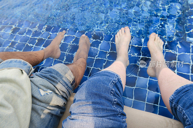 这是一张白人和黑人男子的脚、脚趾、毛茸茸的腿和脚踝在游泳池里溅起水花的照片，种族和谐与平等。一名英国人和印度人坐在游泳池边上，脚和毛茸茸的腿在蓝色马赛克瓷砖下的水里晃荡