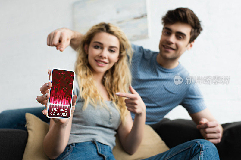 一名快乐的男子和一名金发女子用手指指着智能手机，屏幕上显示着交易课程应用程序
