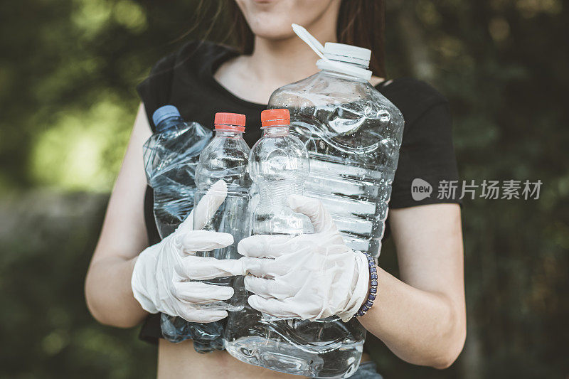 女志愿者穿着衣服在公园里捡垃圾和塑料垃圾。小女孩戴着手套，在户外把垃圾放进黑色的塑料袋里。