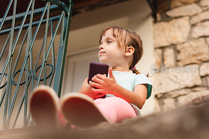 可爱的小女孩坐在楼梯上，拿着手机在户外