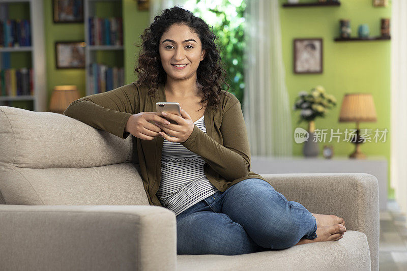 一个年轻女性坐在家里的沙发上用手机发网络信息的照片:-库存照片