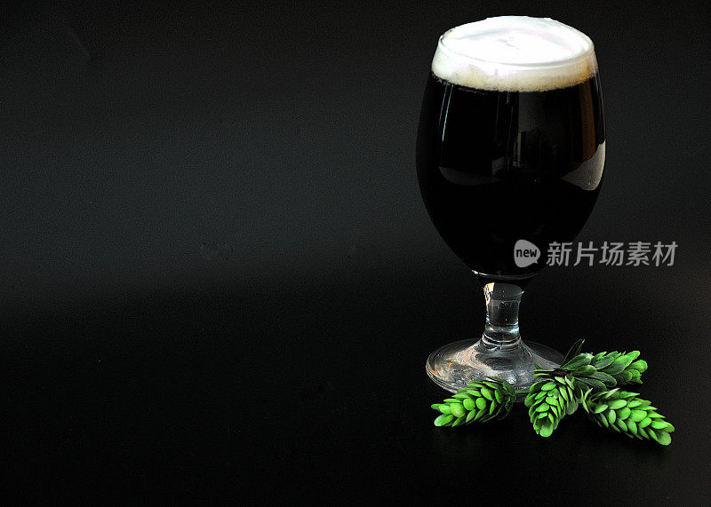 一大杯深色啤酒，白色泡沫和成熟啤酒花在黑色的背景上。