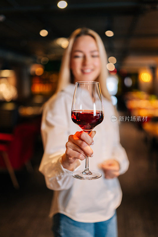 垂直肖像快乐的金发女人拿着一杯红酒站在餐厅与豪华的内部，看向别处。