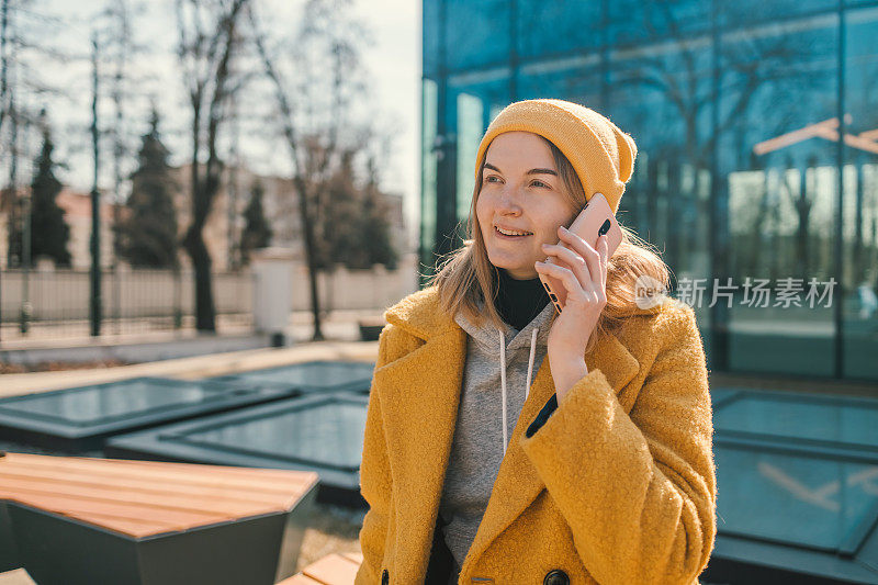 幸福的年轻女子在漫游中打电话，穿着休闲的运动衫和黄色外套，露出了高兴的表情，走在模糊的城市大楼前，享受着智能手机聊天