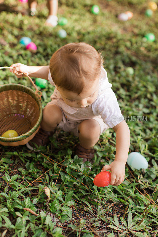 一个可爱的古巴裔美国15个月大的男婴，浅棕色头发和棕色眼睛，穿着棕色兔子和胡萝卜工作服和棕色鞋子，手拿复活节彩蛋篮，享受他的第一次复活节彩蛋寻找庆祝复活节假期