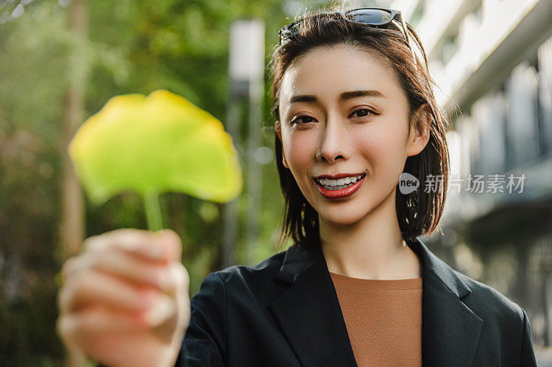 一个迷人的女人站在城市的街道上，手里拿着一片银杏叶。