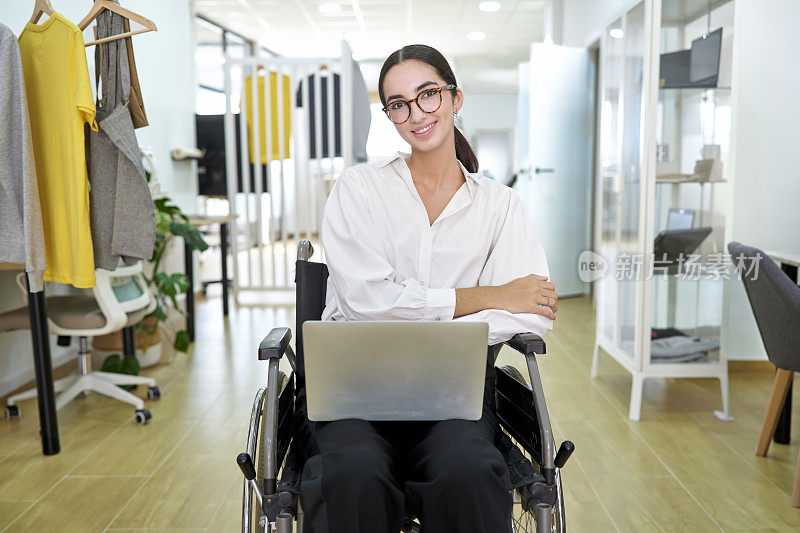 一个漂亮的女孩坐在轮椅上微笑着，在共享办公中心用笔记本电脑工作，摆姿势对着镜头