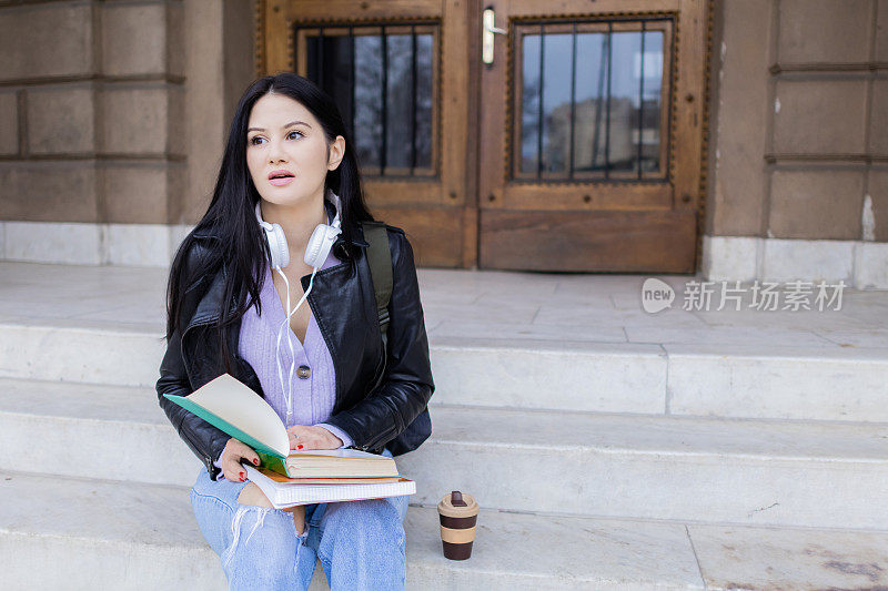 年轻漂亮的女大学生背着双肩包坐在大学楼梯上。她戴着耳机，穿着皮夹克，等待着大学课程的开始。