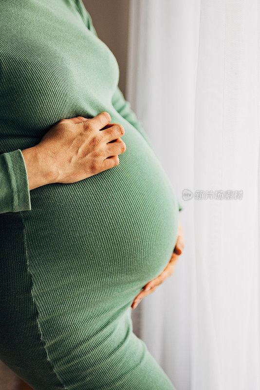 无法辨认的孕妇穿着绿色的衣服，抱着她怀孕的肚子在家里，孕妇，怀孕和母性的概念