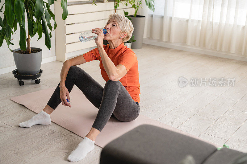 漂亮的老女人运动后喝水。活跃的资深女性在室内练习瑜伽。老年人的运动