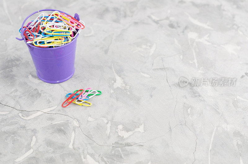 一个装满彩色回形针的紫色桶，另外一堆彩色回形针放在灰色的旧水泥上