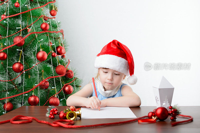 给圣诞老人写信的概念。可爱的小女孩拿着一支笔坐在圣诞树旁的桌子旁