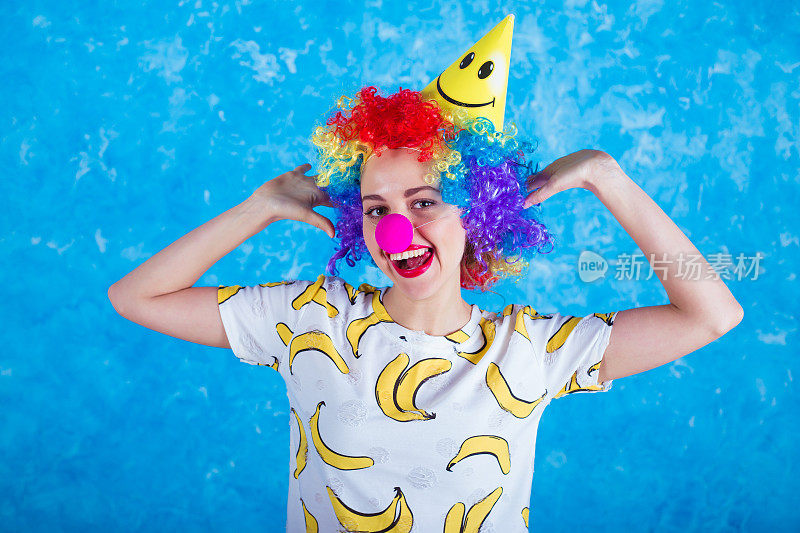 快乐的小丑。假期的概念。一个快乐的年轻女孩穿着小丑服在蓝色背景的相机前摆姿势。