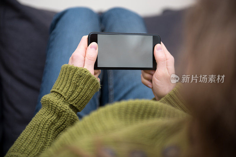 一个金发的年轻女子正坐在家里，手里拿着手机，看着一个空白的智能手机。