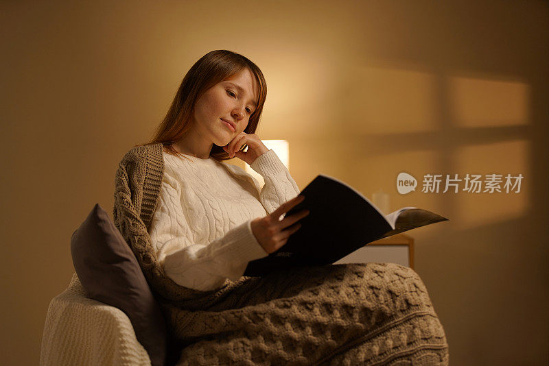 一个漂亮的女孩晚上坐在家里舒适的椅子上看书。梳妆台上有一个小灯，发出温暖的光。