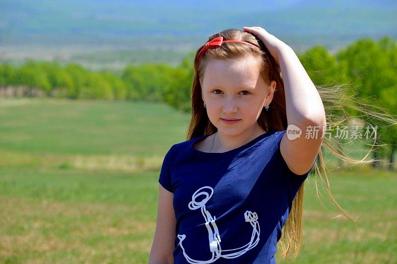 肖像10岁的俄罗斯金发女孩长头发，发带背景的绿色田野。可爱的脸，看着镜头