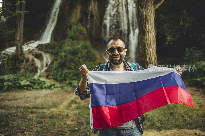 一个带着俄罗斯国旗的瀑布旁边的男游客-一个带着他的国家标志的游客-爱他的国家