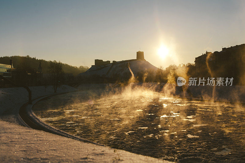 在维尔纽斯寒冷的早晨，水蒸汽在河流和雪岸上升——日出时的第一缕阳光和覆盖河流的薄雾。