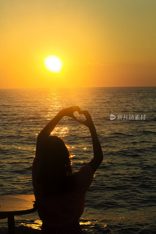落日背景上一个女孩的剪影，握着海边的心型手指