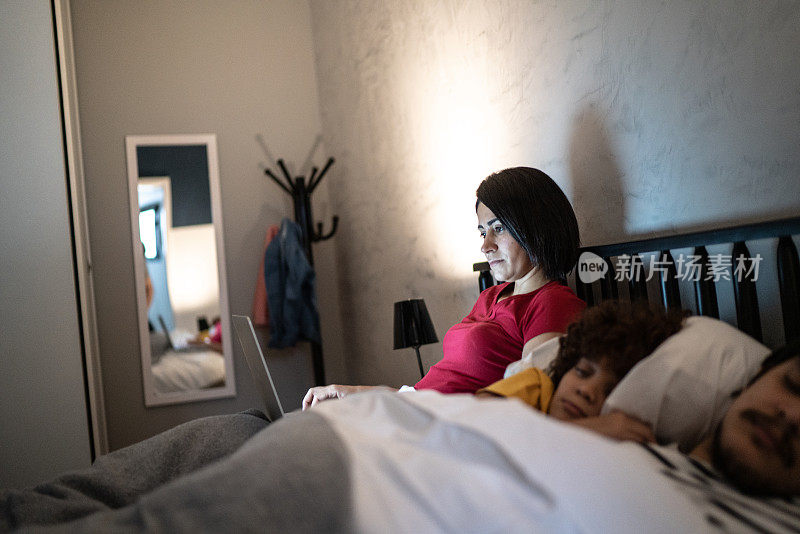 中年妇女在床上使用笔记本电脑，家人睡在她身边
