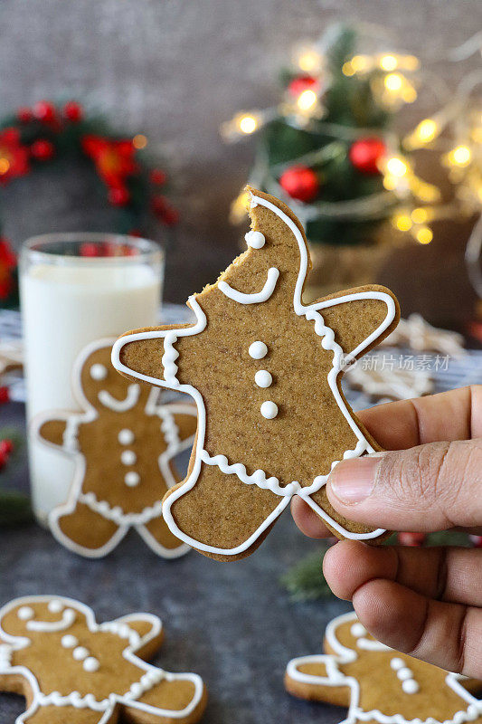 一个无法辨认的人拿着一个姜饼人饼干，没有咬口，用白色的皇家糖霜装饰，喝牛奶，迷你圣诞树，红色和金色的装饰物，花环，灰色的背景，家庭烘焙