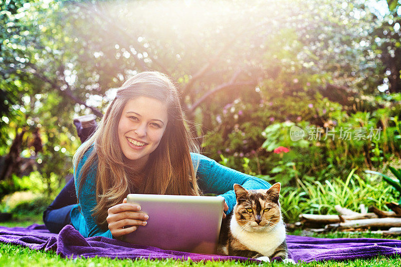 美丽的年轻女子和她的宠物猫躺在户外花园的毯子上，用一个数字平板电脑