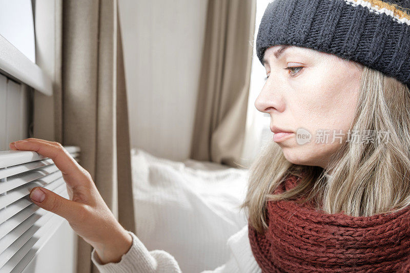 年轻的冷冻妇女穿着毛衣颤抖和冻结的冬天寒冷与震惊的表情在脸上。女孩正在电暖炉上用呼吸暖手。不舒服的呆在家里。