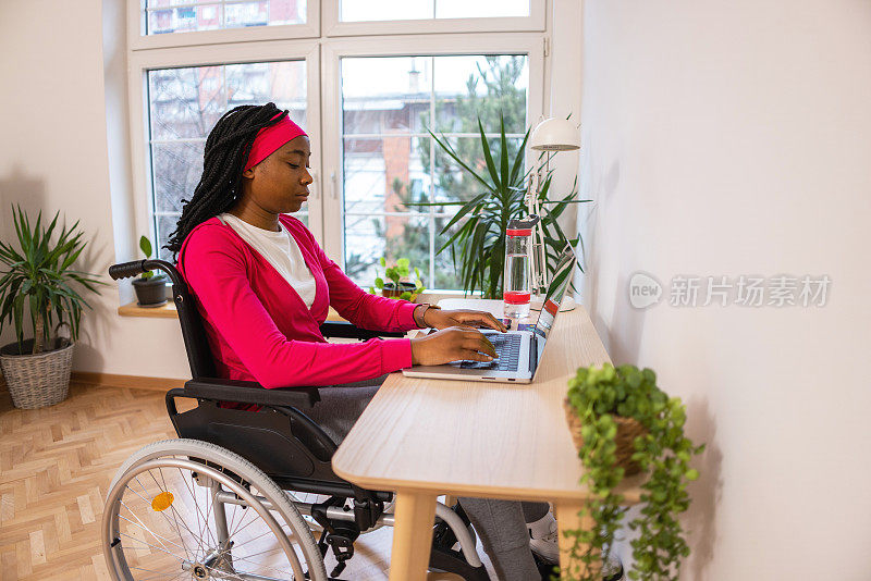 一个黑人妇女坐在轮椅上用笔记本电脑工作的侧面视图，从她的家庭办公室