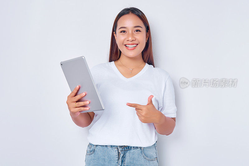 穿着白色t恤的年轻亚洲女孩微笑着指着平板电脑，看着孤立在白色背景上的相机