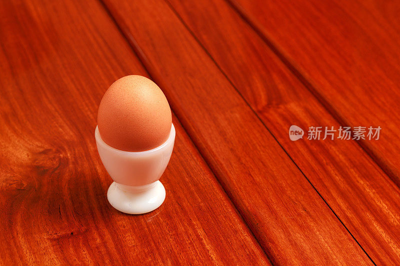 一个鸡蛋在一个白色钢化玻璃蛋杯放置在红木餐桌。副本的空间。没有人