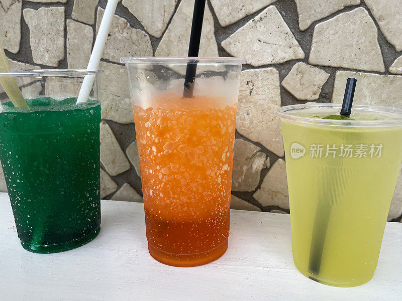一排充满活力的彩色冰冷饮，桌子上有吸管，一次性塑料杯，黄色柠檬水，绿色和橙色冰沙饮料，疯狂的瓷砖墙，重点在前景