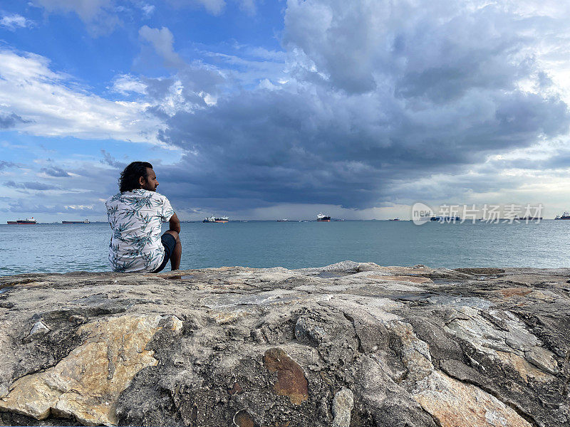 印度男游客度假时坐在岩石上，开阔的大海，波浪，地平线上的油轮，阳光明媚的蓝色多云的天空，新加坡东公园海滩，后视图