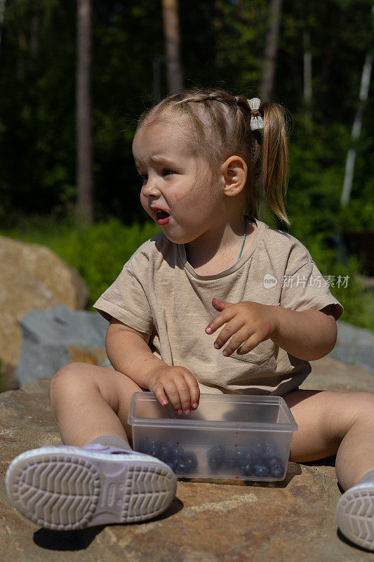 在一个炎热的夏日，一个女孩坐在公园里的大花岗岩上吃草莓。这个女孩吃富含维生素的浆果。孩子在公园散步时吃零食的概念。