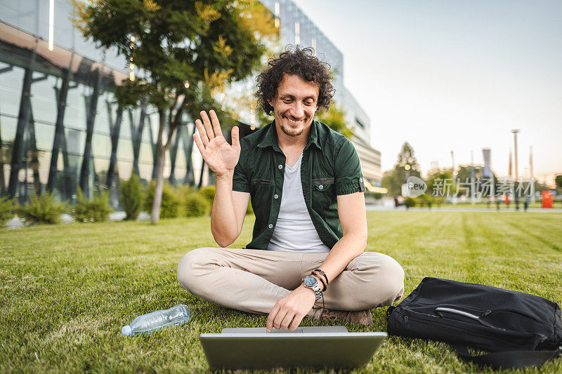 一名男子坐在公园的草地上用笔记本电脑进行视频通话