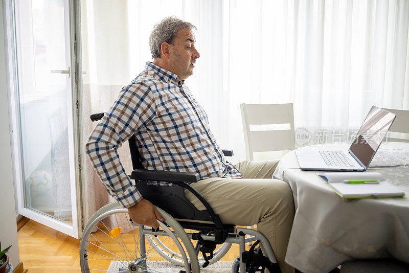 中年残疾高加索人坐在轮椅上，在家工作时用笔记本电脑进行视频通话