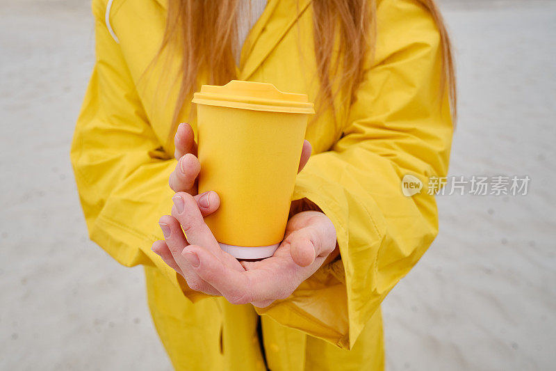 女人手里拿着咖啡的黄色纸杯。是时候在城市里喝咖啡了。咖啡外带。享受这一刻，休息一下。一次性纸杯特写。美味的热饮。空白的文字，模型