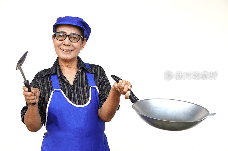 亚洲老年妇女，系着蓝围裙，戴着蓝帽子，拿着煎锅和锅铲或长柄勺。